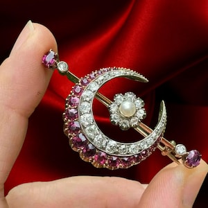 Puede incluir: Un broche de media luna dorada con un centro floral y una barra. La media luna está incrustada con piedras preciosas rosas y diamantes blancos. Una perla se encuentra en el centro de la flor.