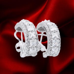 May include: A pair of silver-toned hoop earrings featuring a central row of square-cut clear stones flanked by two rows of smaller round clear stones. The earrings have a hinged closure mechanism and a polished metallic finish throughout the design.