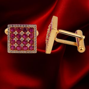 May include: A pair of gold cufflinks with square faces. Each cufflink features a grid of round, ruby-colored stones interspersed with smaller, clear stones. The edges are lined with small, clear stones. The background is a deep red.