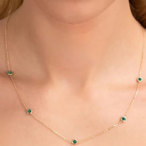 May include: A delicate gold necklace featuring small, round emerald-green gemstones spaced along the chain. The necklace is displayed against a skin-toned background, highlighting the jewelry's design and color.