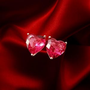 May include: A pair of heart-shaped stud earrings with faceted, ruby-red gemstones. The earrings are set in gold-colored metal and rest on a deep red satin fabric. The gemstones have a brilliant cut, reflecting light.