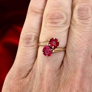 May include: A gold ring featuring two round, vibrant red gemstones. The ring's band is a delicate gold color, and the gemstones are set close together, creating a striking visual contrast against the skin.