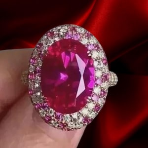 May include: An oval-shaped ring featuring a large, faceted magenta gemstone. The gemstone is surrounded by a halo of smaller pink and clear stones, set in a gold band. The ring is displayed against a deep red background.