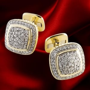 May include: A pair of gold-toned cufflinks with a square design. The center of each cufflink is covered in small, sparkling stones, surrounded by a border of similar stones. The background is a deep red.