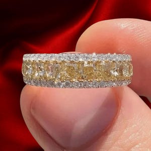 May include: A gold and silver ring with a band of yellow and white gemstones. The ring features a row of yellow gemstones in the centre, bordered by rows of smaller white gemstones. The ring is held up against a red background.