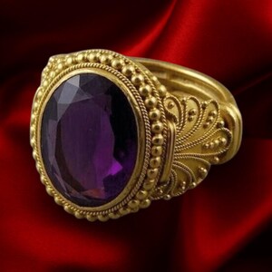 May include: A gold ring featuring a large, oval-cut purple amethyst gemstone at the center. The setting is surrounded by a beaded border and intricate filigree metalwork along the sides of the band, showcasing a detailed, textured gold finish.