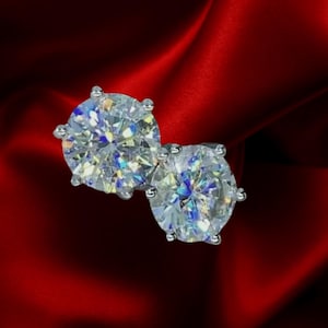 May include: A pair of sparkling diamond stud earrings. Each earring features a large, round, clear gemstone set in a silver or white gold prong setting. The earrings are displayed against a rich, red satin background.