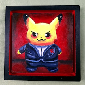 May include: A framed painting of Pikachu wearing a Team Rocket uniform. The yellow character has a stern expression and red cheeks. The dark blue suit has a red "R" emblem. The background is red, and the frame is black.