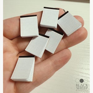 May include: Miniature white notepads with black binding and lined pages are held in a hand. The notepads are rectangular and stacked. The Black Sheep Miniatures logo is in the lower right corner.
