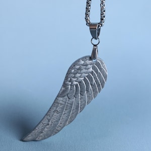 May include: A silver-toned metal pendant shaped like a single bird wing with detailed feather engravings, suspended from a matching silver-toned box chain necklace. The pendant features a textured, weathered surface finish and a polished metal bail.
