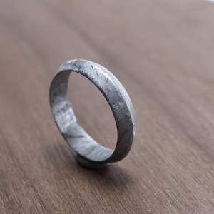 May include: A textured, gray metal ring with a band approximately 0.25 inches wide. The ring has a matte finish and sits on a wooden surface. The ring's surface has a unique, natural pattern.