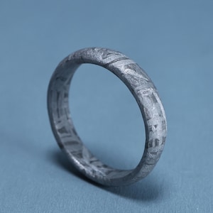 May include: A dark grey metal ring featuring a distinct, etched Widmanstätten pattern across its entire surface. The band has a uniform width and a textured, matte finish that highlights the crystalline structure of the metallic material.