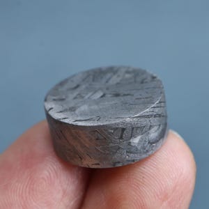 May include: A small, cylindrical iron meteorite specimen featuring a distinct Widmanstätten pattern on its flat, metallic gray surface. The object displays a complex, intersecting crystalline structure with dark gray and silver-toned geometric lines.