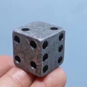 May include: A six-sided die crafted from a dark gray metallic material featuring a distinct, intricate crystalline Widmanstätten pattern across its surfaces. The die has circular, recessed pips arranged in standard numerical configurations.