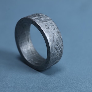 May include: A grey metal ring featuring a distinct, intricate cross-hatched pattern across its outer surface. The band has a flat profile and a matte finish, showcasing a complex geometric texture that covers the entire exterior of the circular ring.
