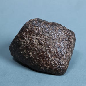 May include: A dark brown, irregularly shaped rock with a rough, pitted, and textured surface. The object features a dense, granular appearance with subtle variations in tone across its uneven, weathered exterior, showcasing a natural, solid form.