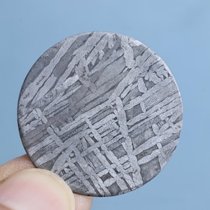 May include: A circular slice of iron meteorite featuring a distinct Widmanstätten pattern of intersecting metallic bands in shades of silver and dark grey. The surface displays a complex, crystalline lattice structure with varied geometric shapes.
