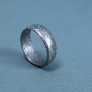 May include: A metallic ring featuring a distinct Widmanstätten pattern across its surface. The band has a polished, cool-toned gray finish with a complex, crystalline, cross-hatched texture that covers the entire exterior of the circular jewelry piece.