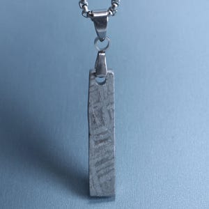 Puede incluir: Un colgante rectangular de tono plateado con una superficie texturizada y grabada, suspendido de una cadena metálica. El colgante tiene una orientación vertical con un pequeño orificio circular en la parte superior para su sujeción.
