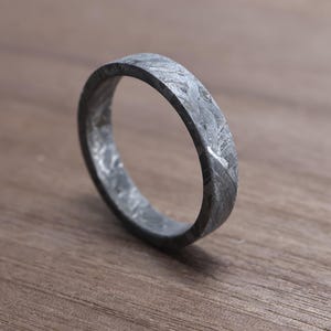 May include: A close-up of a textured, gray ring with a unique pattern. The ring is made of a dark, metallic material and sits on a wooden surface. The ring's design is simple, yet striking.