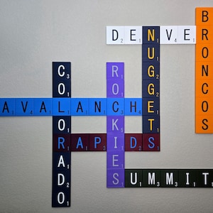 Könnte beinhalten: Ein Kreuzworträtsel aus Scrabble-Steinen zeigt die Namen der Denver-Sportteams: Avalanche, Nuggets, Broncos, Rapids und Summit. Die Steine sind in verschiedenen Farben, darunter Blau, Orange und Lila.