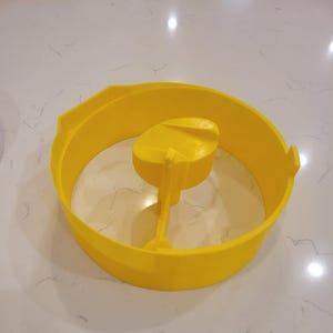 May include: Bright yellow plastic object with a circular outer ring and a central, raised component. The object appears to be a mold or a tool, possibly for crafting or food preparation. The design includes a handle-like extension.