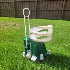 May include: A green and white plastic marble run toy set on a grassy lawn. The toy features a spiral track, a ladder, and several white support pieces. The background includes a wooden fence and green grass.