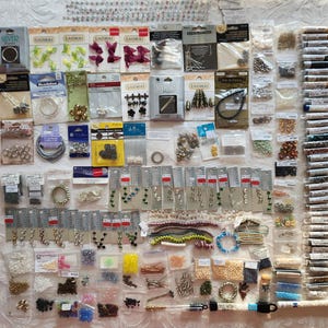 May include: A large collection of assorted jewelry making supplies including various beads, metal findings, clasps, and wire components packaged in individual plastic bags and on cardboard display cards, arranged in a grid-like layout on a surface.