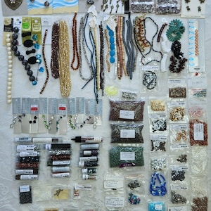 May include: A large collection of assorted jewelry-making supplies including various strands of beads in colors like blue, gold, brown, and black, metal charms, and numerous small plastic bags filled with loose beads, findings, and components.