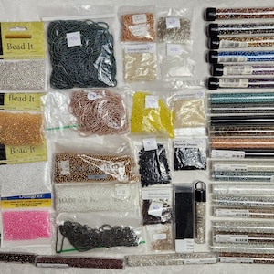 May include: A collection of jewelry-making supplies, including various colors of seed beads in bags and tubes. There are also strands of blue beads, packages labeled "Bead It," and spools of thread. The beads come in a variety of colors, including gold, silver, pink, and black.