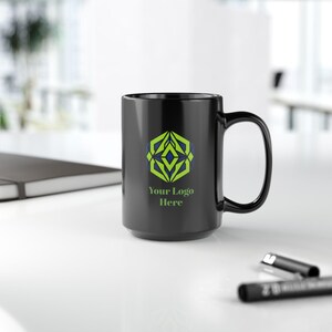 May include: A black ceramic mug featuring a geometric green and blue emblem centred on the front. Below the emblem, the text "Your Logo Here" is printed in a light green font. The mug has a standard handle and a smooth, glossy finish.
