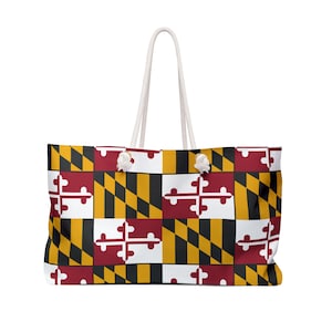 May include: A rectangular tote bag featuring the Maryland state flag pattern in red, white, black, and gold. The bag has two white rope handles attached with silver-toned metal grommets, providing a sturdy grip for carrying personal items.