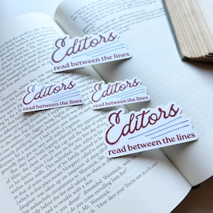 May include: Four stickers with the text "Editors read between the lines" in burgundy script and blue lines, placed on the open pages of a book. The stickers are rectangular.
