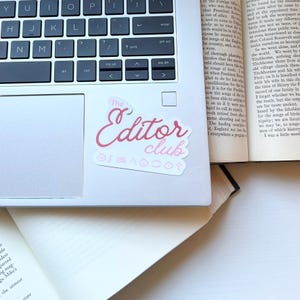 May include: A pink sticker with the words "The Editor Club" in a cursive font is affixed to a laptop. The sticker also features several small symbols below the text. The laptop keyboard is visible.