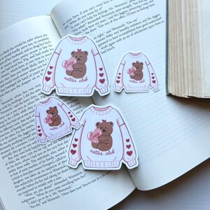 May include: White sticker featuring a brown teddy bear holding a pink heart with the word "Cozy" and "editor club" text. The sticker has pink heart accents and is on a book page.