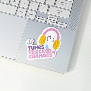 May include: A sticker with a pink and yellow headphone design and musical notes, with the text "TUNES & TRACKED CHANGES" in pink and purple. The sticker is placed on a silver laptop.