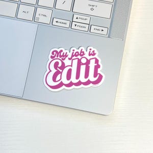 May include: A sticker with the text "My job is Edit" in a retro, bubble-style font. The text is in a vibrant pink color with a white outline, placed on a silver laptop. The sticker is likely for editors or those in media.