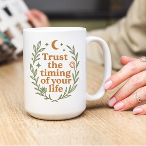 Puede incluir: Taza de cerámica blanca con asa, con el texto "Trust the timing of your life" en marrón estilizado. El texto está rodeado por una corona de hojas verdes, flores y estrellas. La taza está sobre una superficie de madera marrón claro.