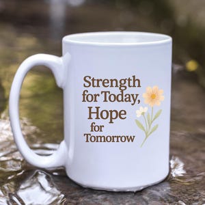 Puede incluir: Taza de cerámica blanca con asa curva. La taza presenta el texto "Strength for Today, Hope for Tomorrow" en marrón, junto con una ilustración floral. La taza está sobre un fondo borroso al aire libre.