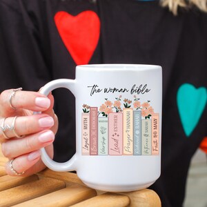 Puede incluir: Taza de cerámica blanca con el texto "The Woman Bible" y detalles florales. La taza presenta un diseño de libros apilados con palabras inspiradoras. La taza es sostenida por una persona que lleva una camisa negra con forma de corazón.