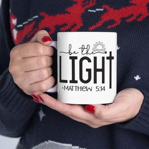 Puede incluir: Taza de cerámica blanca con el texto "be the LIGHT" en escritura negra y letras de bloque, con un diseño de cruz. La taza también presenta un gráfico de sol y olas y el texto "MATTHEW 5:14".