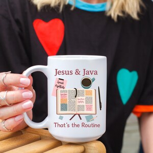Puede incluir: Taza de café de cerámica blanca con el texto "Jesus & Java" y "That's the Routine". La taza presenta una ilustración de una taza de café, un libro abierto y un bolígrafo. La taza la sostiene una persona que lleva una camiseta negra con diseños de corazón.