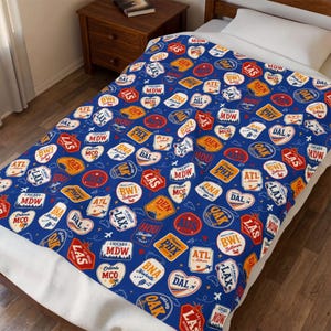 May include: A blue throw blanket featuring a repeating pattern of various airport code labels in red, orange, and white. Each label displays a three-letter city code such as MDW, LAX, ATL, DEN, and HOU, along with city names and small airplane icons.