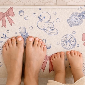 Op de afbeelding: Crèmekleurige badmat met een grillig ontwerp met een badeend, zeep, bellen en strikken in roze en blauw. Voeten van volwassenen en kinderen zijn zichtbaar op de mat, wat duidt op een gezinsvriendelijk badkameraccessoire.