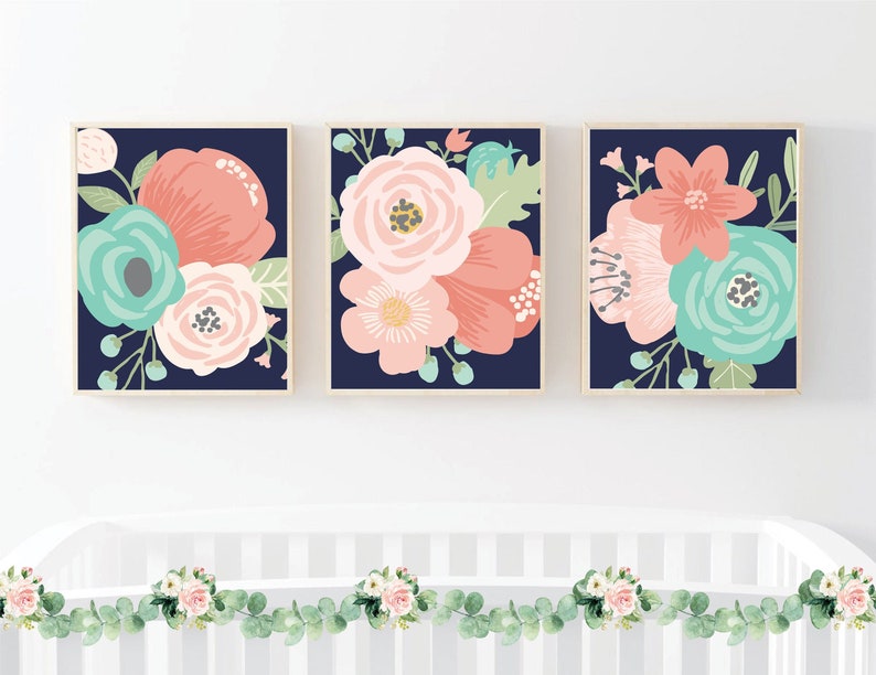 May include: Three framed floral prints with pink, peach, and teal flowers on a navy blue background. The prints are hanging on a white wall above a white crib with a green and white garland.
