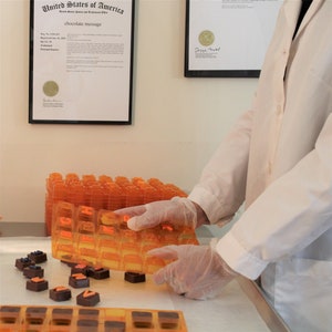 May include: A person wearing white gloves is holding a clear orange plastic mold with letters for making chocolate. The mold is filled with dark chocolate squares. There are other molds on the table.