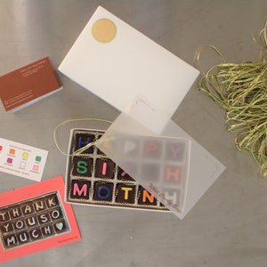 Puede incluir: Una caja blanca con una tapa de pl&aacute;stico transparente que contiene un juego de letras de chocolate que deletrea "HAPPY SIXTH MOM". La caja est&aacute; sobre una superficie gris con una tarjeta rosa que dice "THANK YOU SO MUCH" y una peque&ntilde;a tarjeta de visita naranja.