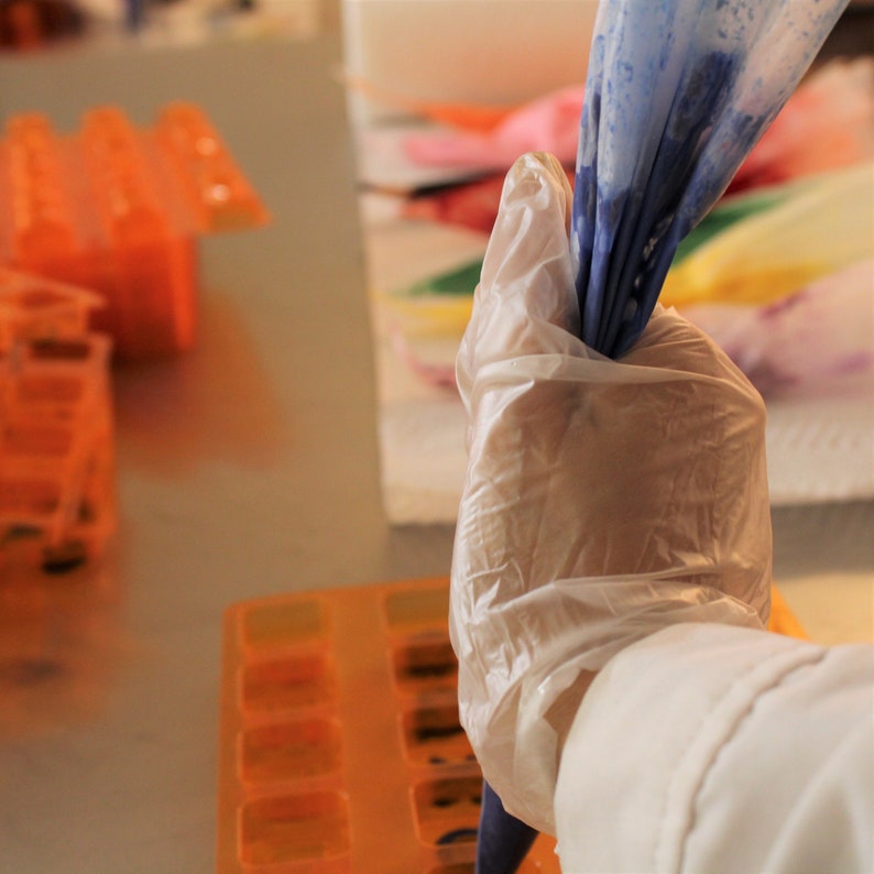 May include: A gloved hand holds a blue piping bag filled with frosting. The bag is being squeezed to release the frosting. The hand is wearing a white glove. The background is out of focus and shows a tray of orange silicone molds.