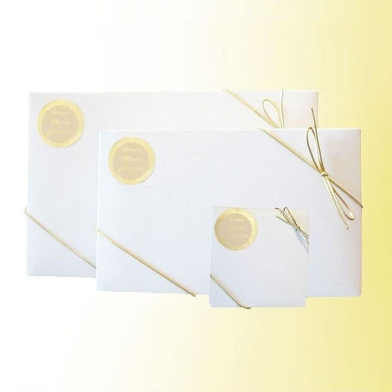 Puede incluir: Tres cajas de regalo blancas de diferentes tama&ntilde;os, atadas con una cinta fina de color marr&oacute;n claro. Cada caja tiene una pegatina redonda beige con letras doradas.