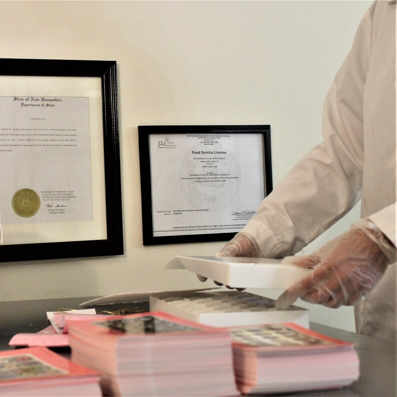 May include: A white framed certificate with the text "Food Service License" and a gold seal. The certificate is hanging on a white wall next to a framed document. A person wearing white gloves is holding a white box with a stack of pink brochures on a table.